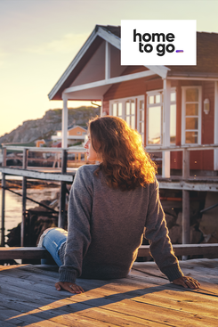 Ferienhäuser buchen über HomeToGo Frau sitzt entspannt vor einem Ferienhaus am See in Skandinavien symbolisiert Angebote von HomeToGo